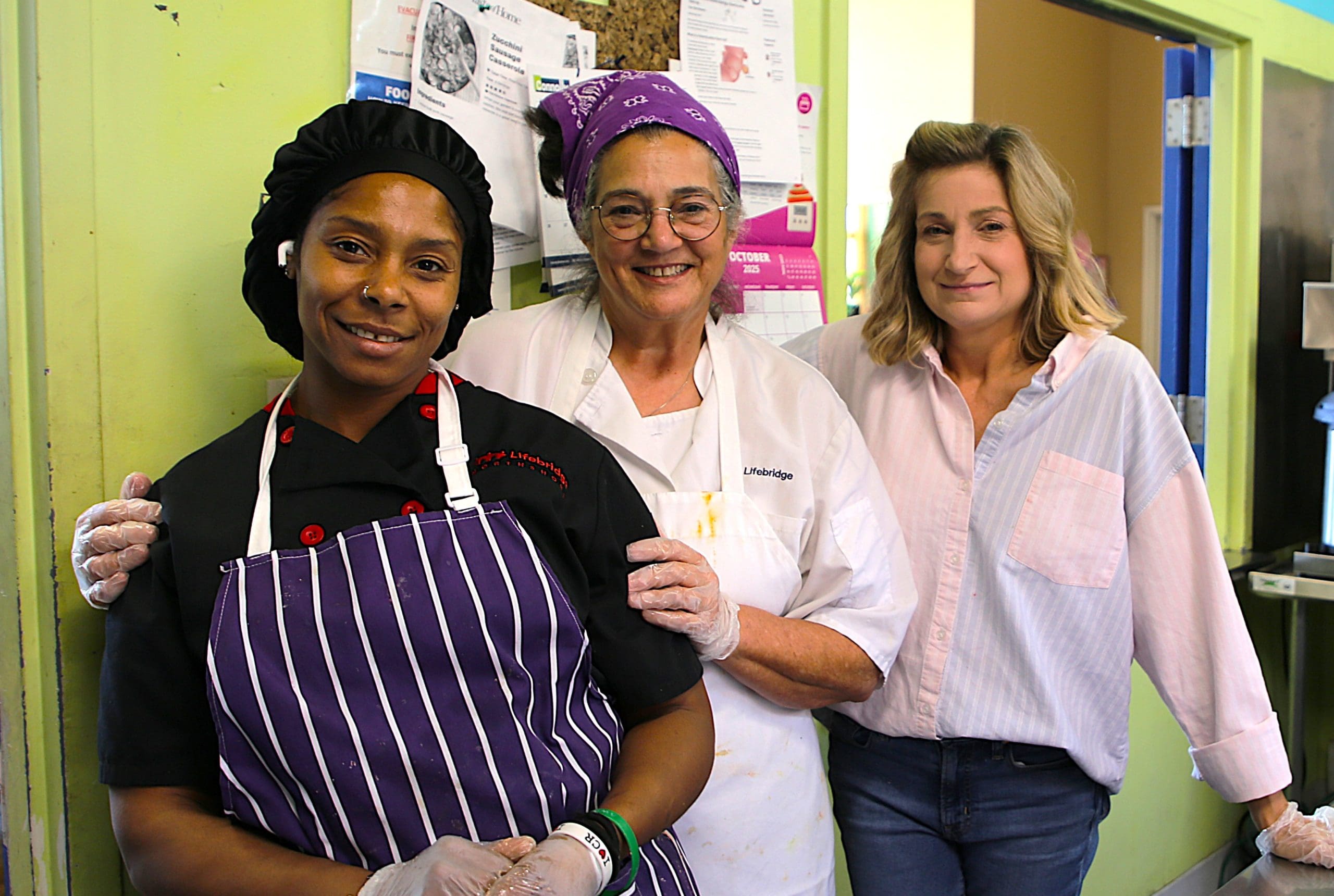 Three women smiling and posing for a picture. Two of the women are wearing chef coats and aprons, the other is wearing jeans and a button down shirt.
