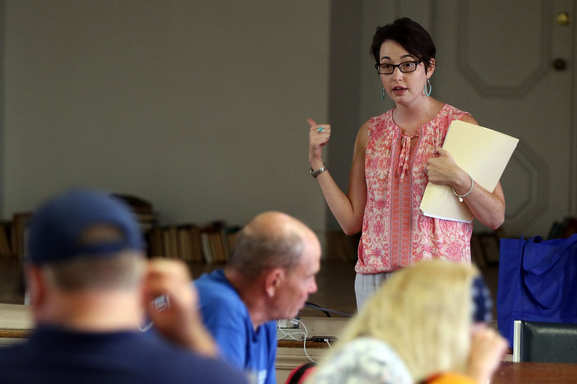A photograph of a woman holding a folder speaking to a group of people.