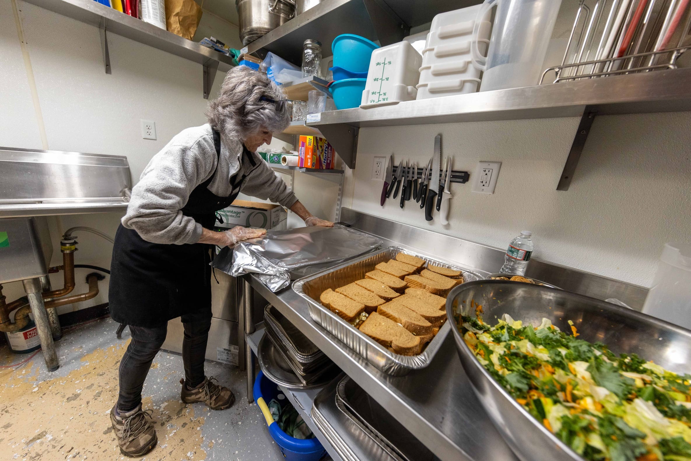 A woman in an apron covers pans of sandwiches with tin foil.