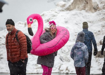 Woman with pink sunglasses in a pink flamingo floaty smiling at the camera with people walking by.