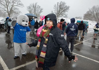 A man in a winter coat poses for the camera while someone in a seal mascot costume waves in the background.