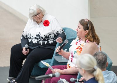Two women sitting in arm chairs, one speaking into a microphone and the other looking at her.