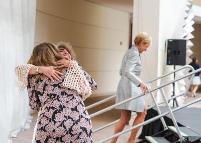 Two women hugging. A third woman in the background is smiling and walking up the steps to a stage.