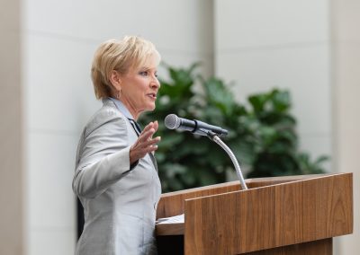 A woman is speaking at a podium while gesturing to the right.