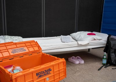 A cot with a pillow, blanket, and notebook laid on it. There is a backpack and orange "Rentacrate" leaning on the cot. A pair of pink slides are under the cot.