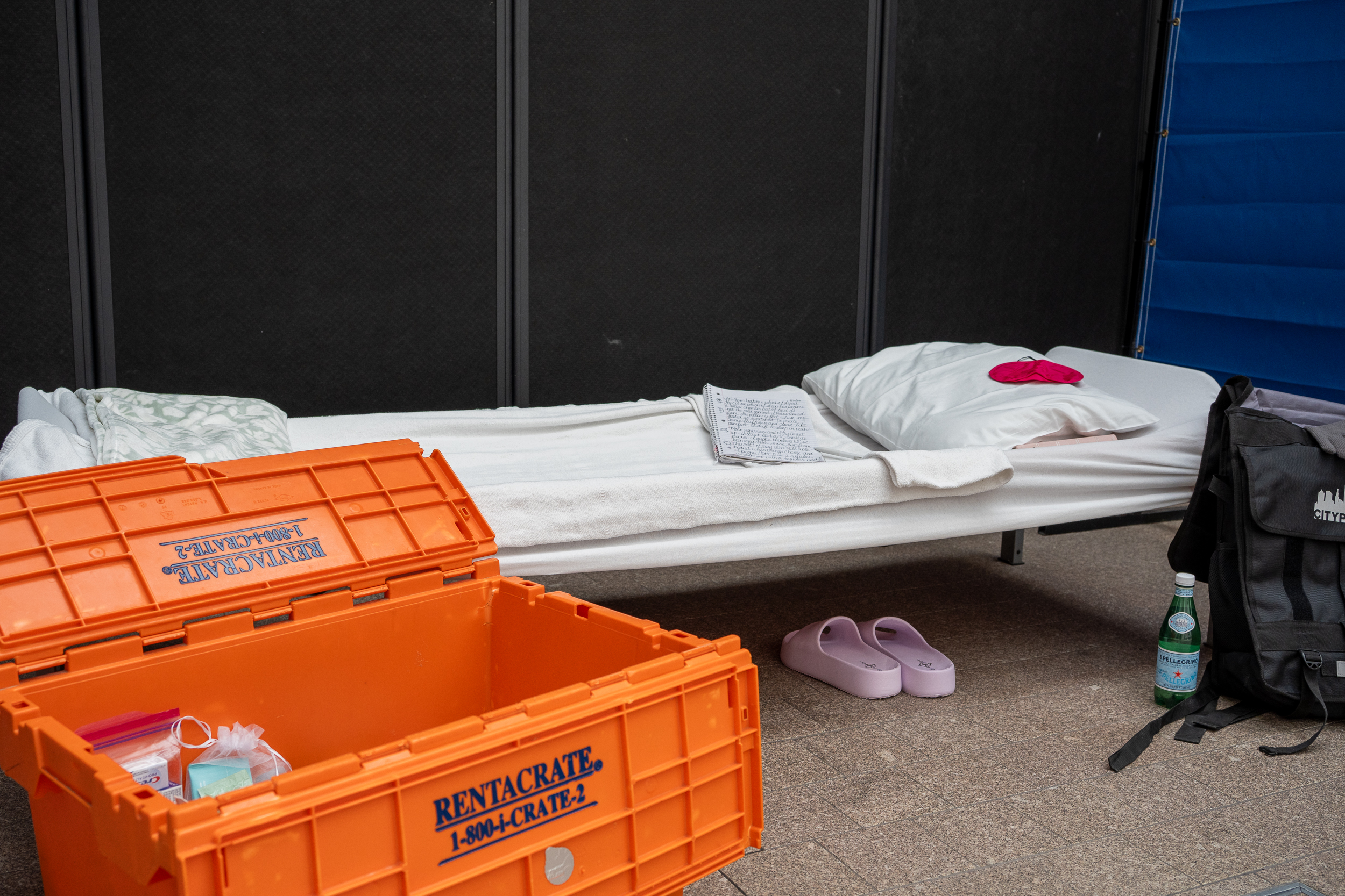 A cot with a pillow, blanket, and notebook laid on it. There is a backpack and orange "Rentacrate" leaning on the cot. A pair of pink slides are under the cot.