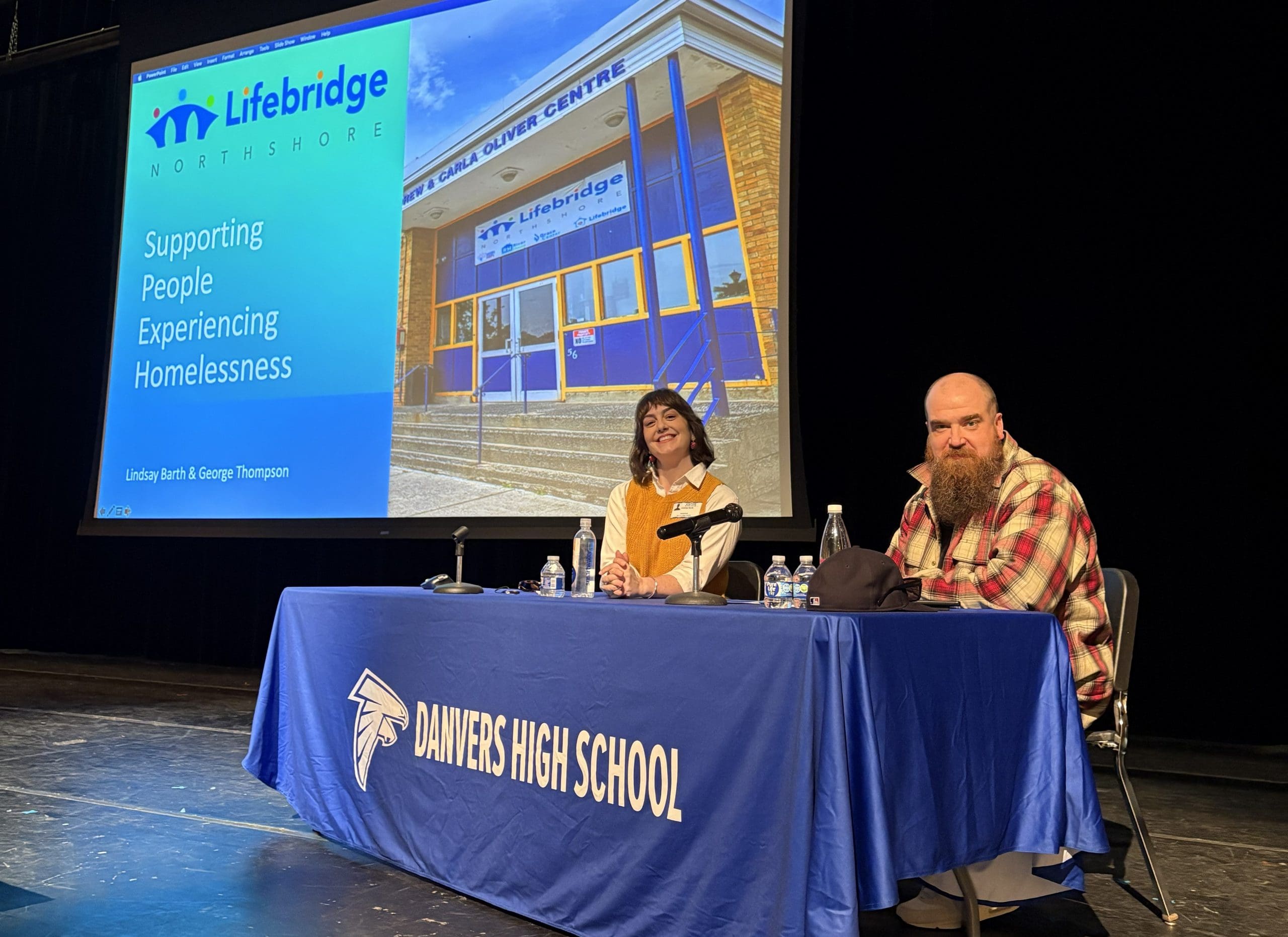 Lindsay and George sit behind a table with a tablecloth that says "Danvers High School." They are on a stage in front of screen showing a photo of Lifebridge that says "Supporting People Experiencing Homelessness"