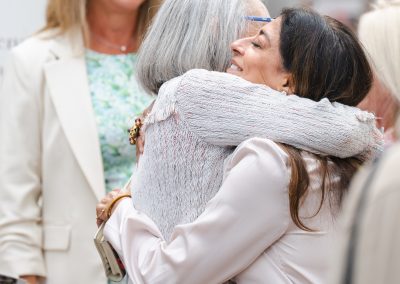The image captures a warm, candid moment of two women embracing at an indoor event, with other attendees visible in the background.