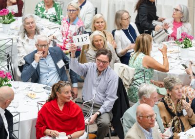 This image shows a similar formal charity luncheon, benefit, or live auction event. Multiple round banquet tables are filled with guests dressed in smart-casual and business attire.