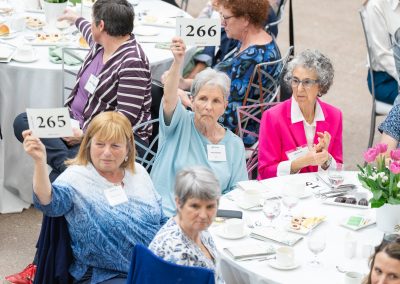 This is a third photograph from the same formal charity luncheon, benefit, or live auction event. Multiple round banquet tables are filled with guests dressed in smart-casual and colorful daywear attire.