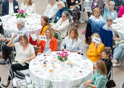 This is a fourth photograph from the same formal charity luncheon, benefit, or live auction event, taken from an elevated wide angle. It captures multiple round banquet tables with guests actively participating in a paddle-raise fundraiser.