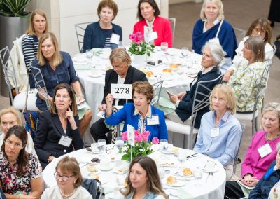 This is a fifth photograph from the same formal charity luncheon or live auction fundraiser, captured from an elevated angle. The image shows several round banquet tables filled with seated guests.