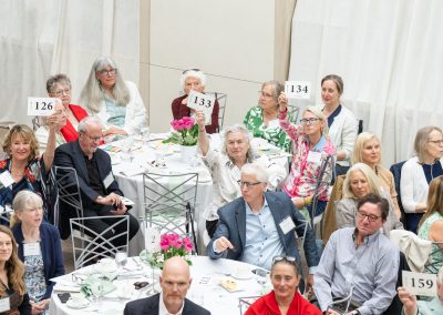 This is a sixth photograph from the formal charity luncheon or live auction fundraising event, captured from an elevated perspective. The image shows multiple rows of round banquet tables packed with engaged attendees.