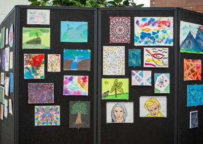 The photograph showcases a vibrant public art exhibition pinned to a dark grey or black multi-panel, freestanding partition wall. The display features a diverse collection of small, multi-colored paintings and drawings, likely created by community members or students.