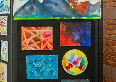The photograph provides a close-up view of a single panel section of the black partition display wall from the ongoing public art exhibition. This specific panel showcases seven distinct, colorful art pieces created by community members or students, utilizing a variety of abstract and landscape styles.