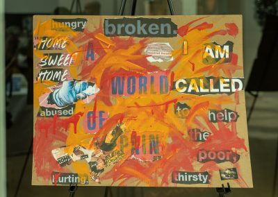 The photograph displays a square mixed-media collage on a brown cardboard backing, resting on a black metal easel stand. The artwork features abstract paint strokes combined with cutout magazine words and imagery to highlight themes of suffering, poverty, and social advocacy