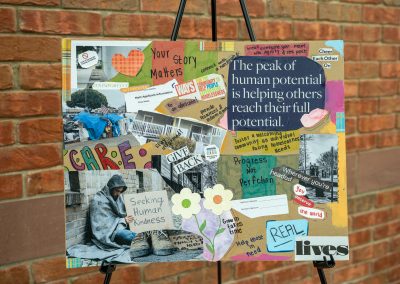 The photograph displays a wide rectangular mixed-media collage poster mounted on a black metal artist easel. Crafted on a sturdy cardboard or brown paper backing, the artwork combines cut-out text, colored paper patterns, handwritten notes, photographs, and magazine imagery to promote themes of empathy, advocacy, and community support for individuals experiencing homelessness