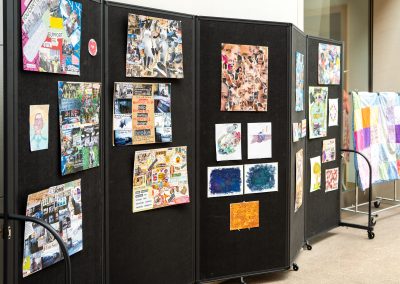 The photograph displays a public art and community project exhibition pinned across a long, black multi-panel folding partition wall on wheels. The panels showcase an array of creative collages, drawings, and text-based art pieces. To the far right, a colorful patchwork quilt stands on a rolling garment rack.