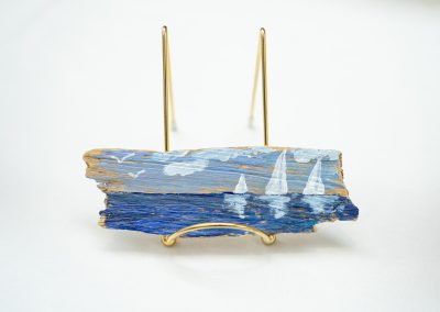 The photograph provides a straight-on close-up view of a unique, small landscape painting created on a rustic piece of weathered driftwood [image_iL_LYP.png]. The art piece rests securely on a polished gold wire display stand against a clean white backdrop
