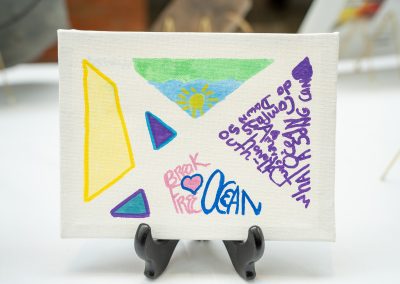 The photograph provides a straight-on close-up view of a small, rectangular canvas painting resting on a decorative black display stand. The artwork features a collection of geometric shapes, simple landscape illustrations, and hand-written text fragments rendered in markers and paint