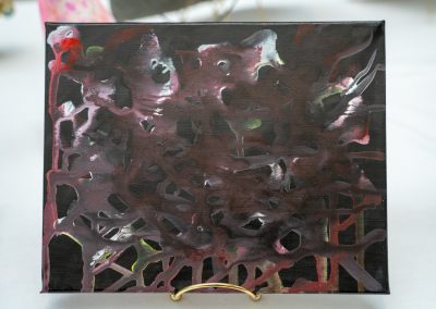 The photograph provides a close-up view of a small, rectangular canvas painting resting on a gold wire display stand. The artwork features a dark, heavily layered abstract composition with an organic, textured appearance
