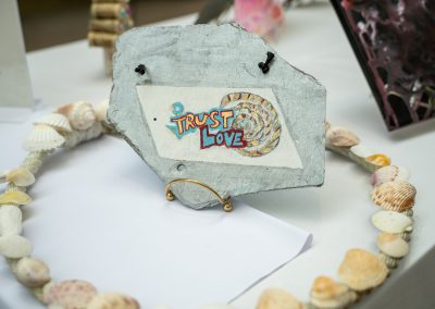 The photograph provides a close-up, angled view of a unique, handcrafted art piece painted on a rough slab of natural stone. The piece rests on a gold wire stand, surrounded by a circular shell-adorned rope wreath on a white tabletop display
