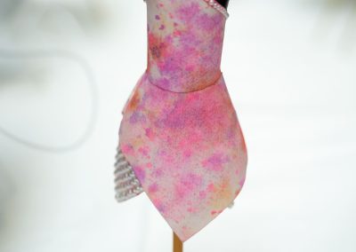 The photograph provides a straight-on, close-up view of a miniature dress form display, showcasing a tiny, hand-designed fashion piece [image_DCpvmm.png]. The delicate sculpture rests on a tabletop surface, blending fabric crafting with miniature sculpture work