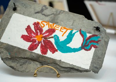 The photograph provides an angled, close-up view of a unique, handcrafted art piece painted on a rough slab of natural stone. The finished work rests on a small gold stand on a white tabletop display surface
