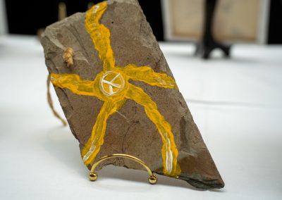 The photograph provides an angled, close-up view of a unique, handcrafted art piece painted on a rough slab of natural stone [image_14rUPU.png]. The piece rests on a small gold stand on a white tabletop display surface
