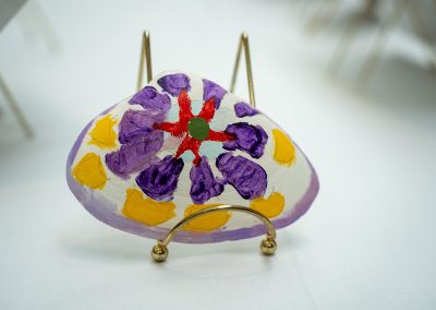 The photograph provides a straight-on close-up view of a small, decorative art piece hand-painted onto a natural clam shell. The piece rests on a polished gold wire stand against a clean white backdrop.