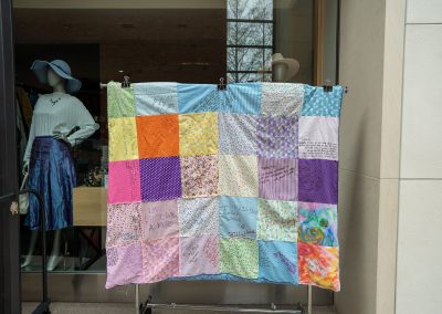The photograph provides a straight-on view of a colorful patchwork quilt hung for display on a rolling metal garment rack. The quilt is stitched together from a variety of patterned fabric squares, many of which contain personal handwritten messages, names, and illustrations from community members.
