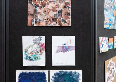 The photograph provides a straight-on, close-up view of a vertical panel section belonging to a black, fabric-covered multi-panel folding partition wall. This specific section features six community or student art pieces neatly arranged and pinned to the surface, showing a mix of collages, illustrations, and abstract textures.