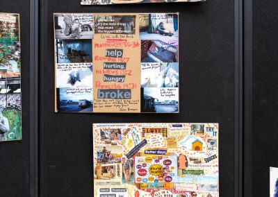 The photograph provides a straight-on close-up view of a vertical panel section belonging to a black, fabric-covered multi-panel folding partition wall. This section features three large, multi-colored mixed-media collages arranged vertically and pinned securely to the surface, focusing heavily on themes of community, faith, hardship, and mental health.