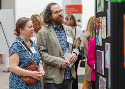 The image shows a candid indoor photograph of a man and a woman standing side-by-side as they look at an art display during a public event or reception.