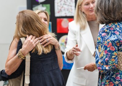 The image shows a candid indoor photograph capturing a warm greeting between several women at a social gathering or gallery event.