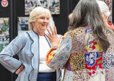 The image shows a candid indoor photograph capturing a lively conversation among three senior women at a social gathering or gallery event.