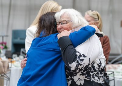 The image shows a candid indoor photograph capturing a joyful and affectionate hug between two senior women at an organized event or reception.