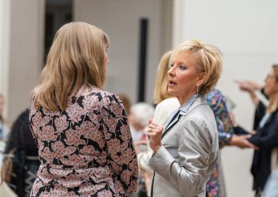 The image shows a candid indoor photograph capturing a conversation between two women at a busy social event or reception.