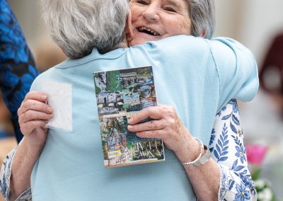 The image captures a joyful, candid moment of two senior women tightly embracing at an indoor social gathering or reception.