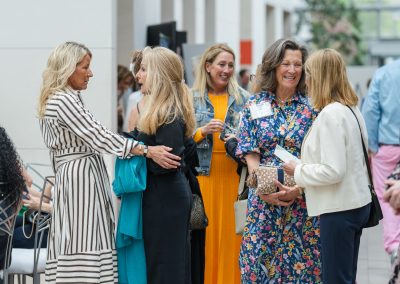 The image shows a candid indoor photograph capturing a large group of elegantly dressed women socializing and conversing in a bright, open atrium during an event or reception.