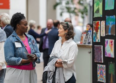 The image shows a candid indoor photograph capturing a conversation between two women at an art exhibition or reception, with other event attendees mingling in the background.