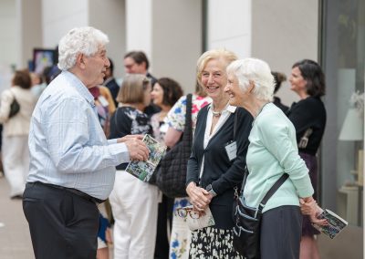 The photograph captures a lively, cheerful interaction among three senior individuals socializing at a crowded indoor event or reception.
