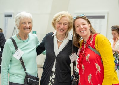 This image features three women standing closely together, smiling brightly for a portrait at an indoor event. They are dressed in colorful, casual-chic attire.