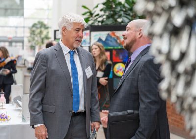 The photograph captures a candid interaction between two professionally dressed men conversing at an indoor event, surrounded by art displays and other attendees.