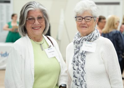 This image shows two women standing side-by-side and smiling for a photo at a busy indoor eventBoth women are wearing white event name tags pinned to their tops, featuring a blue corporate or organizational logo.
