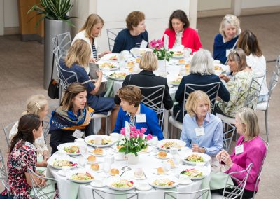 The photograph provides a high-angle, birds-eye view of a formal luncheon or banquet event taking place in a large, modern atrium with tiled flooring. The scene centers on two large, round dining tables where groups of well-dressed women are seated, eating salad and conversing.