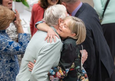The photograph captures an affectionate, joyful embrace between a man and a woman in the center of a crowded indoor event or reception.