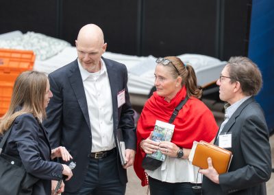 The photograph provides an overhead, high-angle view of a group of four professionally dressed individuals engaged in a serious conversation at an indoor event, standing in front of what appears to be a mock shelter or facility display.