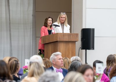 The photograph captures a formal moment during an indoor banquet or luncheon, focusing on two women presenting together at a wooden podium in front of a seated audience