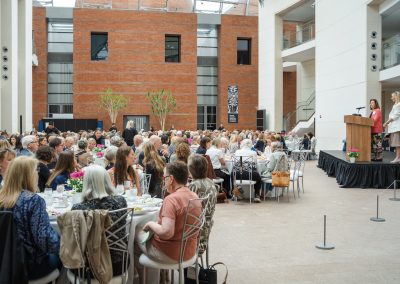 The photograph provides a wide, expansive view of a large formal luncheon or charity banquet event being held inside a massive, modern museum atrium filled with natural light. Dozens of well-dressed attendees are seated at large round dining tables while two female presenters speak from a raised stage on the right side of the hall.